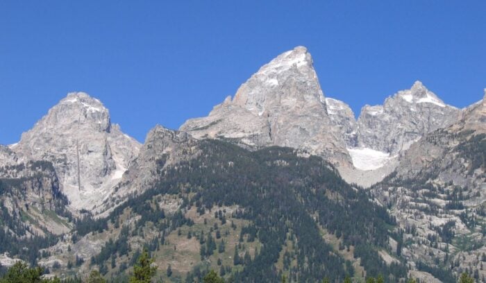 A section of a mountain range with some spots of snow and fir trees in the foreground.