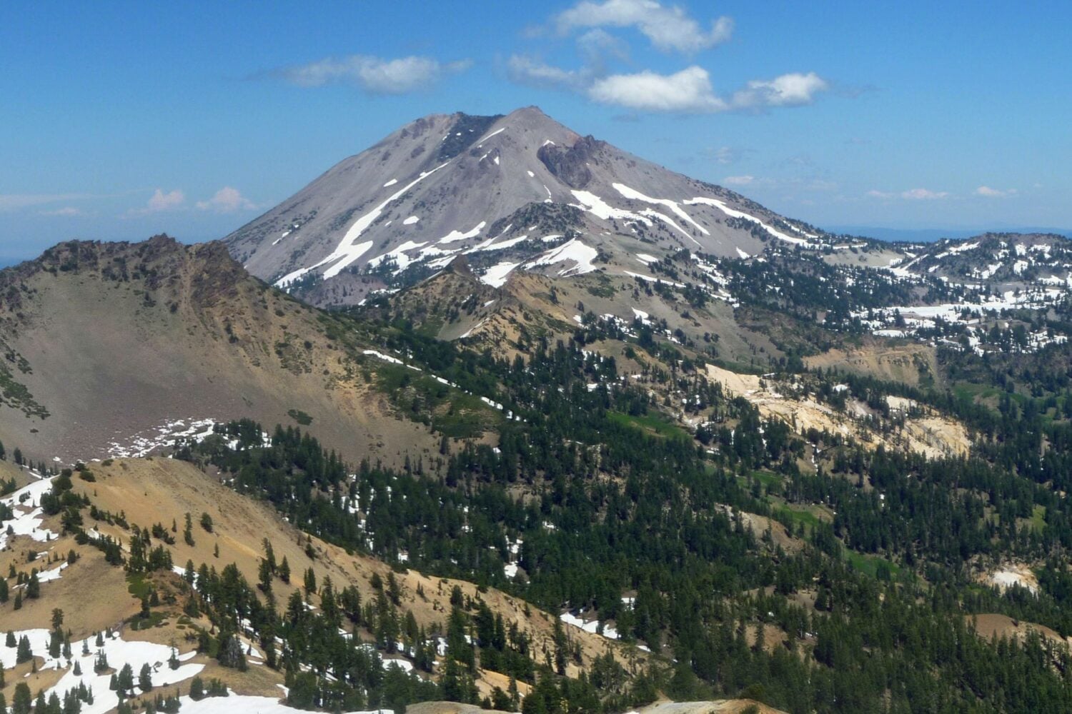 A view across a valley of a dome volcano on a bright sunny day with a blue sky and some clouds.