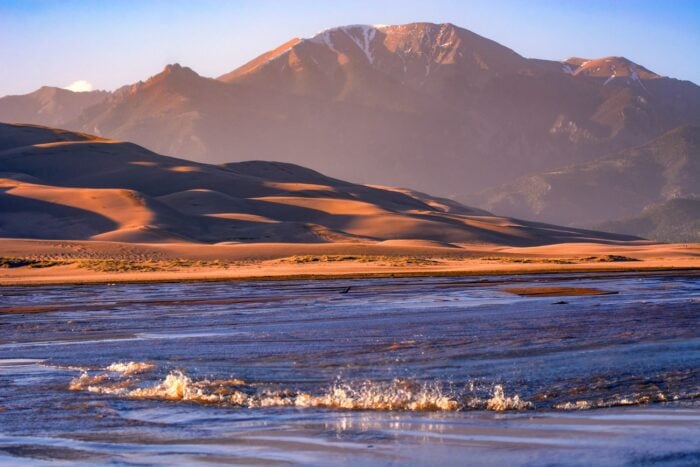 A view of a surge flow on a creek with sand dunes and mountains in the background.